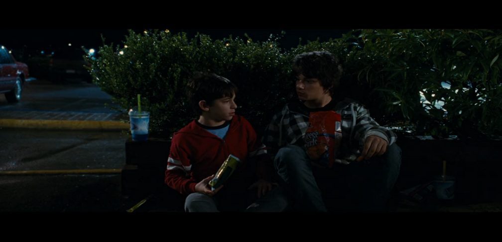 Greg and Rodrick (Devon Bostick), a tan white boy with messy dark hair, sitting outside behind some shrubbery. They are eating snack foods and drinking slushies.