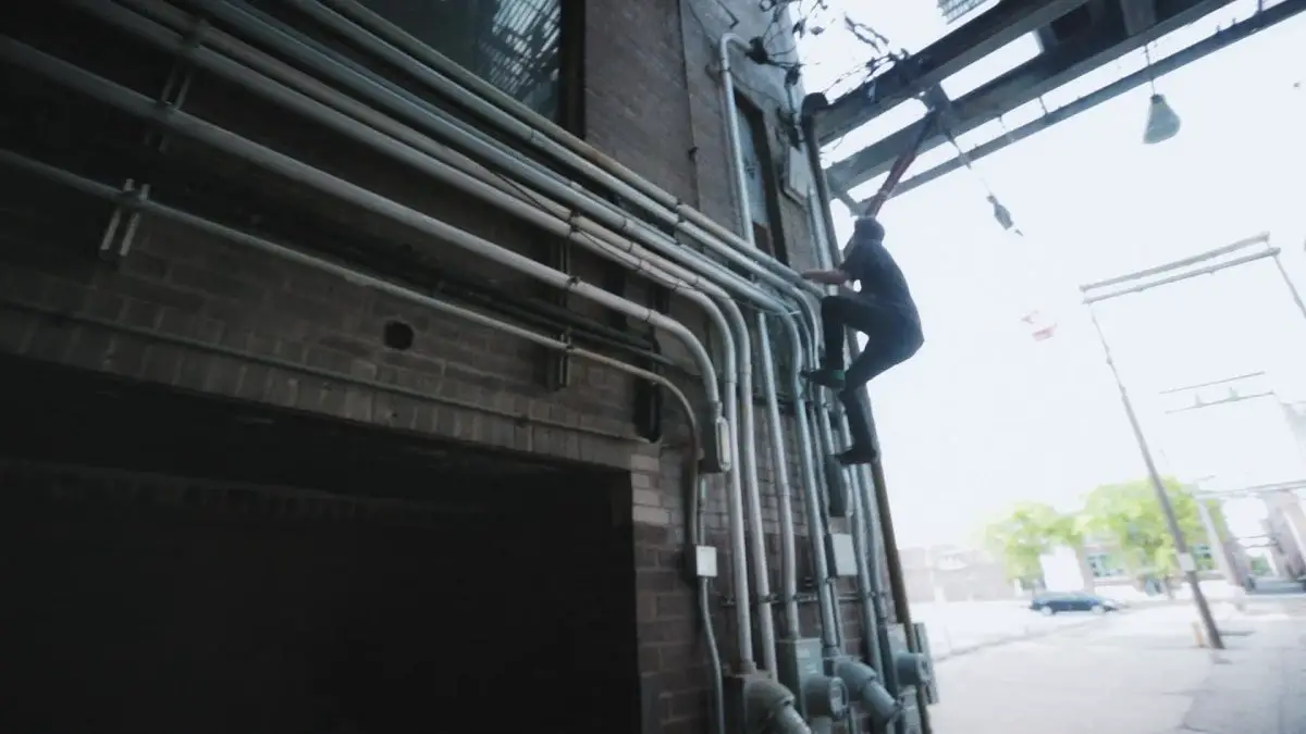 Image from Minding the Gap: A young man dressed in black climbs a set of pipes outside a parking garage.