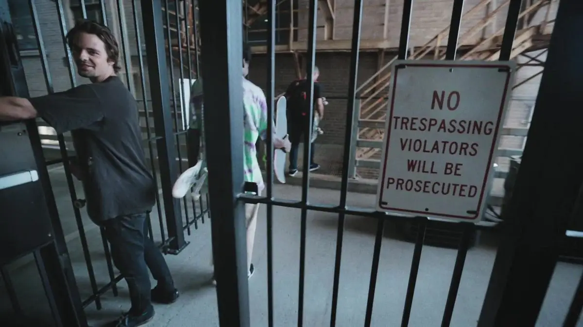 Image from Minding the Gap: A smiling young white man (Zack Mulligan) holds open a gate with a sign saying "No Trespassing Violators Will Be Prosecuted"