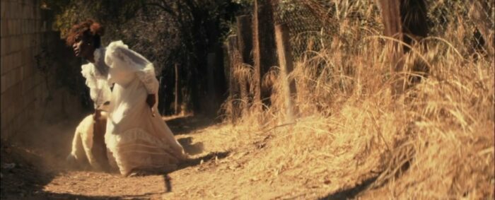 Still from I Dream of You Dream of Me. A woman in a wedding dress drags a fur coat and bag behind her down a dusty path.