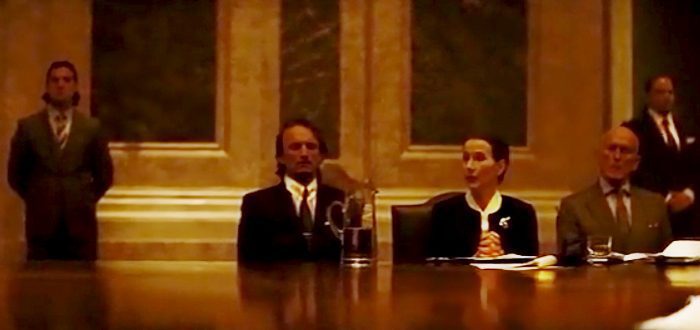 D. Vogel (Brigitte Millar) sits at a large table while addressing a meeting held by Blofeld