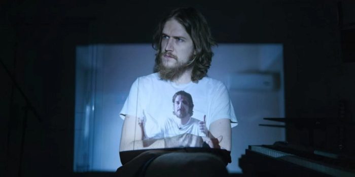 Bo Burnham, a white man with unkempt brown hair and beard, wearing a white t-shirt. He is sitting down in a dark room, a projection of himself playing overtop him.