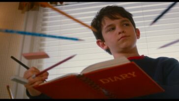 Greg Heffley (Zachary Gordon), a young white kid with dark hair wearing a dark blue shirt, sitting at a glass table. The camera is looking up from underneath the table; we see Greg's red diary open across the table, a pencil in his hand. Several other pencils and an eraser are arranged to be pointing at his head.