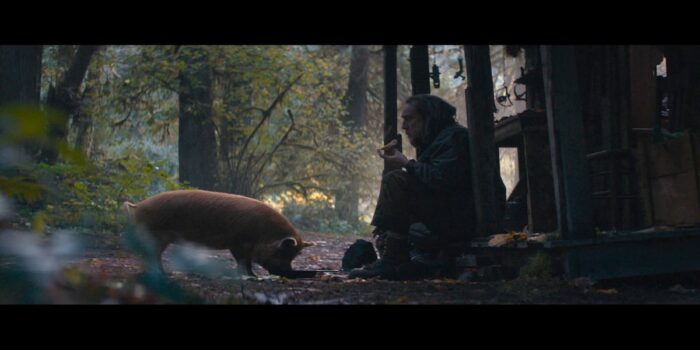 Robin (Nicolas Cage) and his pig outside his home in the forest. Robin is sitting on his shallow wooden porch while the pig stands before him. They are each enjoying a snack.