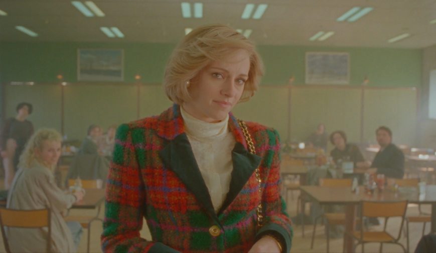 Diana Spencer (Kristen Stewart) in 'Spencer' (2021) standing in a small diner wearing a plaid fuzzy button-up sweater/blazer and a white turtleneck underneath. Her light blonde hair is swept to the left side of the frame. The green-toned dining area is misty, and all the eyes of the customers are on Diana. She looks somewhat awkward or worried.