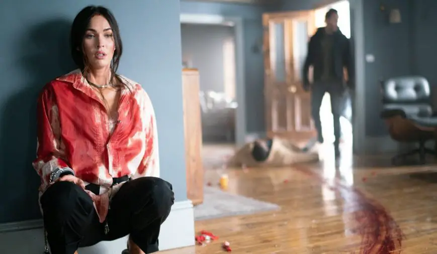 Emma (Megan Fox) against a robin's egg blue wall, her white button-up shirt covered in blood. There is a smear of blood going past her, and red and white rose petals litter the polished wood floor. In the background, a blurry man is entering through the front door.