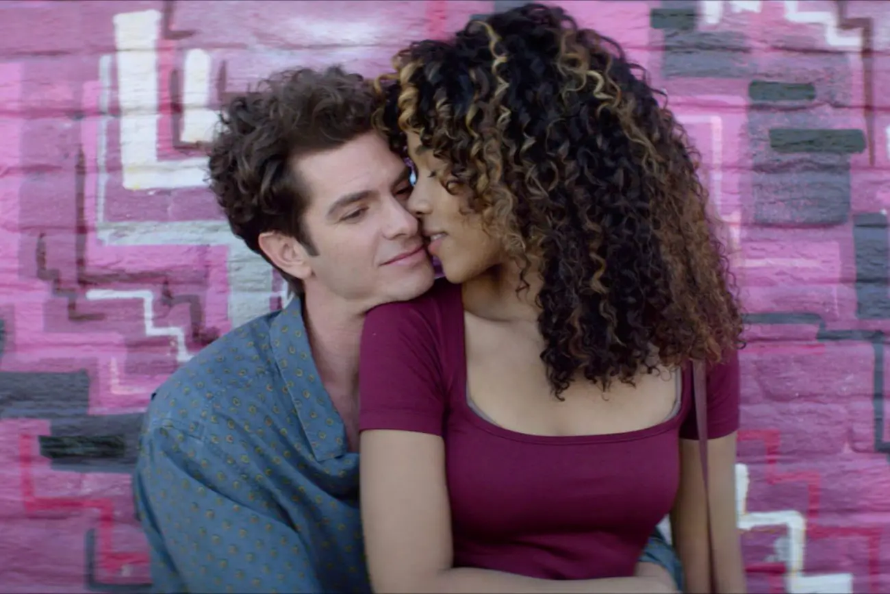 Jon (Andrew Garfield) and Susan (Alexandra Shipp) snuggling up next to each other in front of a brightly-painted brick wall.