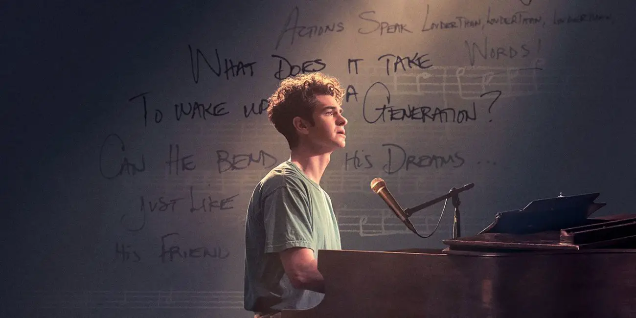 Jonathan Larson (Andrew Garfield) sitting at the piano with a spotlight shining on him, superimposed against a backdrop of music notes and lyric fragments.