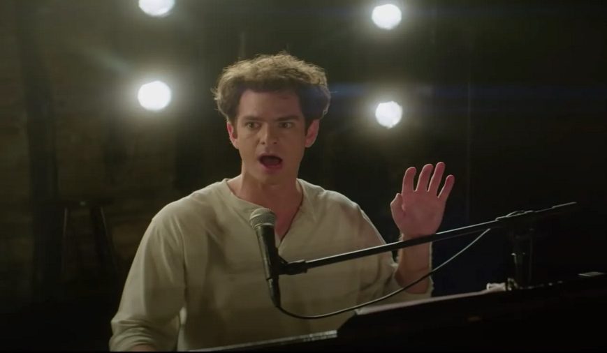 Jonathan Larson (Andrew Garfield) in tick, tick... BOOM! (2021) sitting at a piano on a stage while he is doing a performance. He's looking towards the audience while holding up his left hand. Four lights are behind him and he's singing into a microphone.