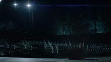 Jonathan Larson (Andrew Garfield) playing a black piano in the lower right corner of the frame. He is outside in some kind of stadium, hundreds of empty seats behind him. Two spotlights are shining down on him, and the bare trees in the background seem to be lit in blue from the ground up.
