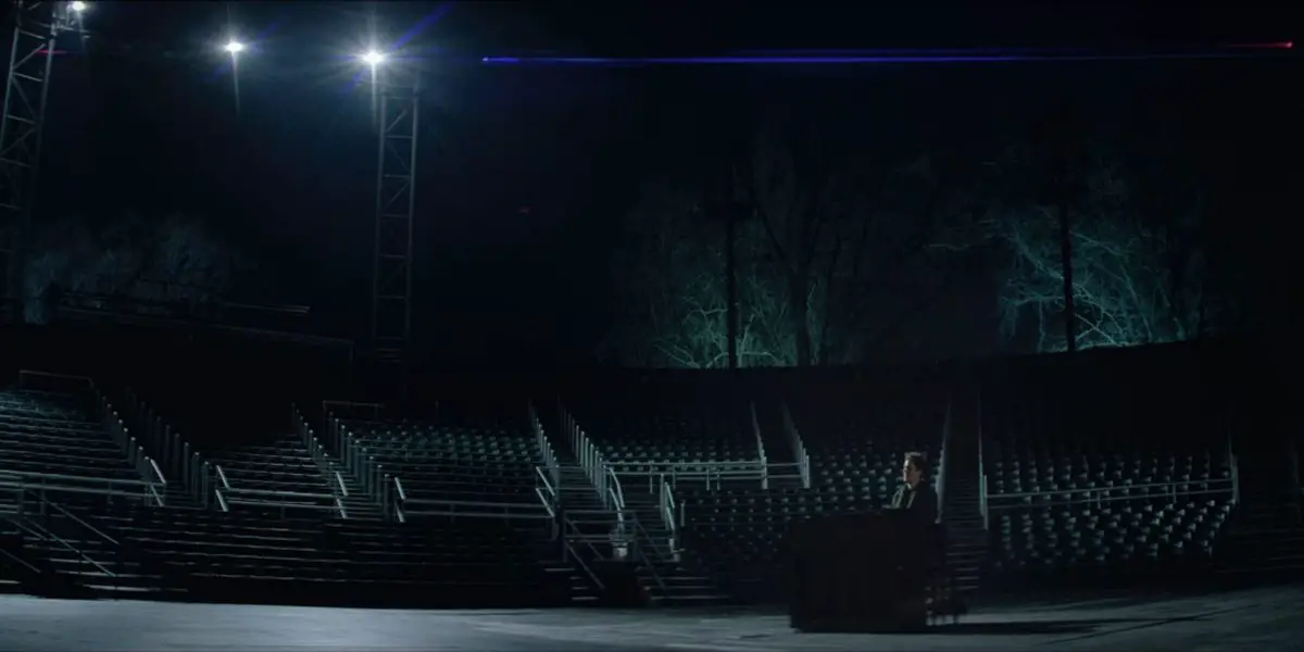 Jonathan Larson (Andrew Garfield) playing a black piano in the lower right corner of the frame. He is outside in some kind of stadium, hundreds of empty seats behind him. Two spotlights are shining down on him, and the bare trees in the background seem to be lit in blue from the ground up.