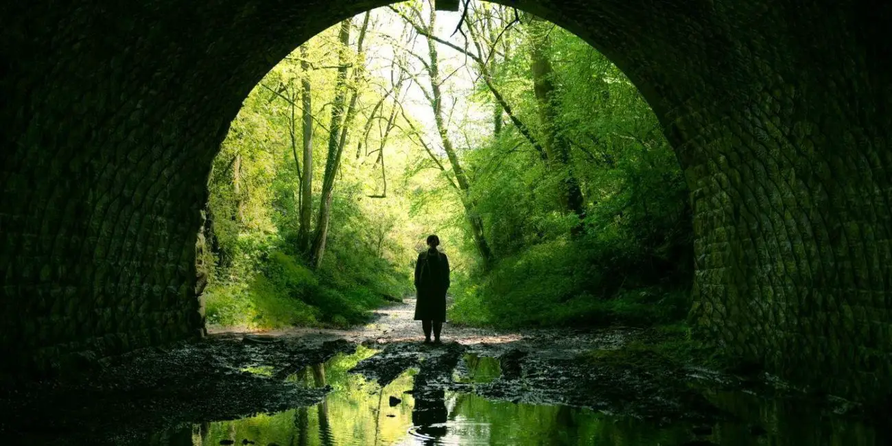 Jessie Buckley (presumably) stands in a green forest at the entrance to a large brick tunnel in the film Men