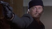 Paul, in a jacket and stocking cap, aims a gun.