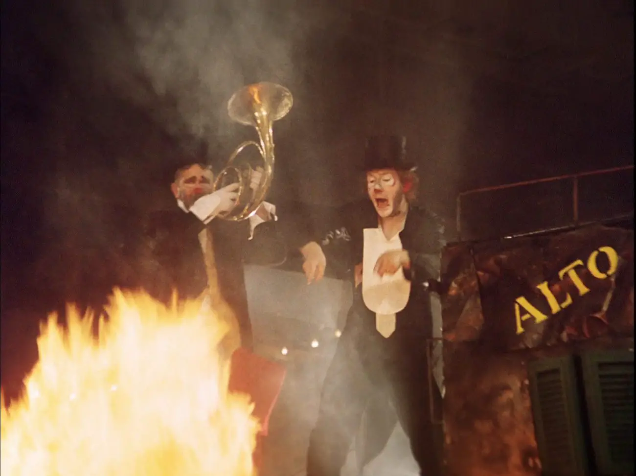 Two clowns standing in front of a fire, one holding a musical instrument