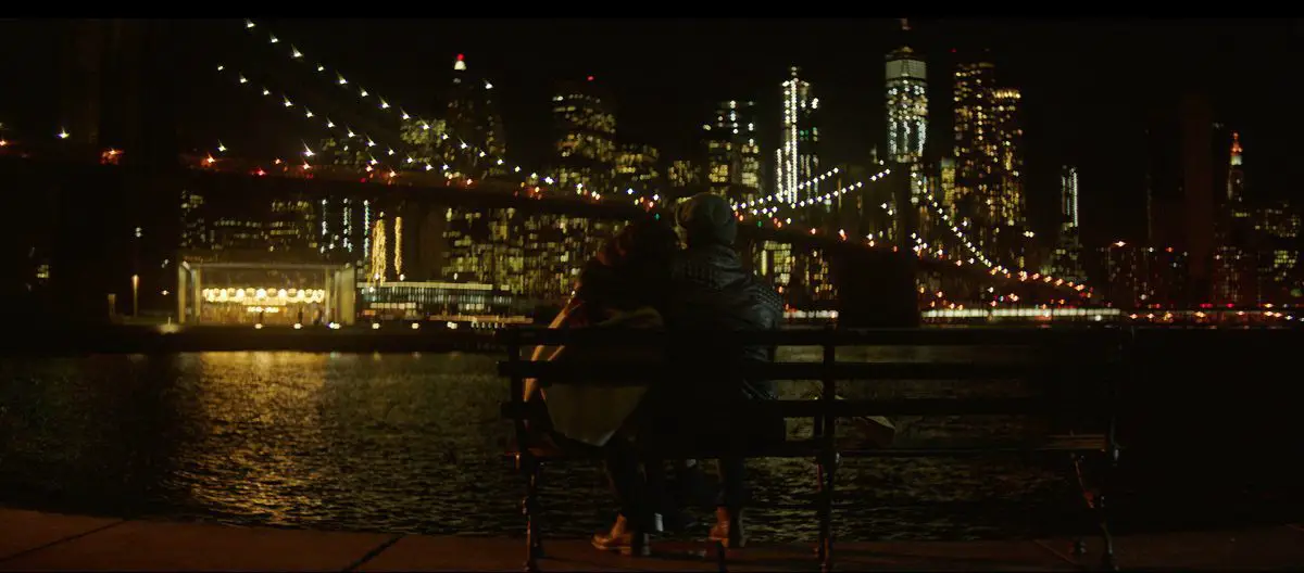 Image from In A New York Minute: A couple sits in front of the Brooklyn Bridge at nighttime.