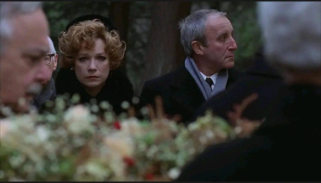 A medium shot of Chance (Peter Sellers) and Eve (Shirley MacLaine) standing in the background of a flower-decorated casket in the foreground of the shot.