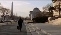 A medium shot of Chance (Peter Sellers) wandering the sidewalks of Washington D.C. with the Washington Monument visible in the distance.