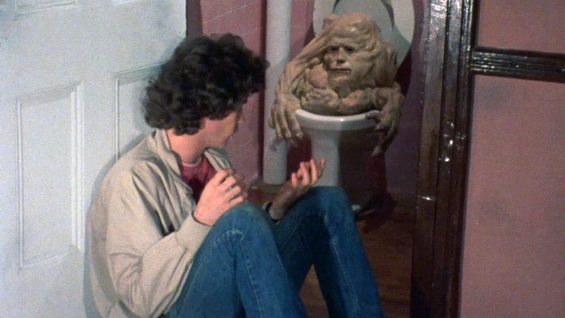 An image from Basket Case shows Duane (Kevin Van Hentenryck) sitting with his back against an open bathroom door, looking to Belial, a fleshy blob with a human face and deformed hands, who is sitting atop an open toilet bowl