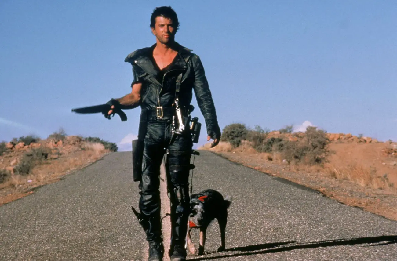 A screenshot from Mad Max 2 shows Max (Mel Gibson) walking down a desolate wasteland road toward the camera with his dog, a sawn off shotgun in his right hand