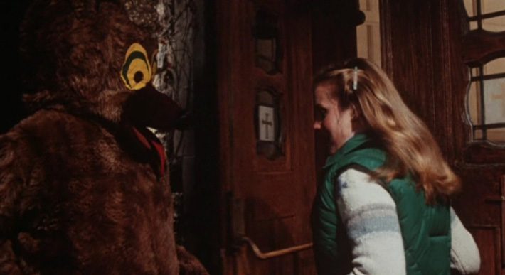 A screenshot from Girls Nite Out shows a girl in a varsity jacket running toward a stoic figure in a mascot bear costume 
