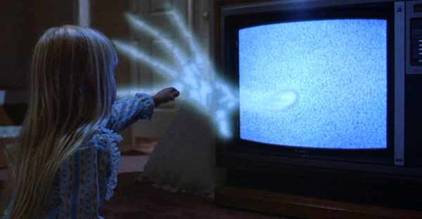 An image from Poltergeist shows a spectral hand reaching out of TV static in a dark room toward Carol Anne (Heather O’Rourke)