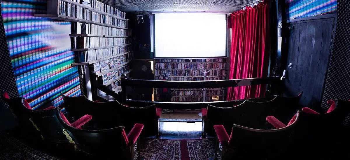 A photograph of the inside of one of 20th Century Flicks’ cinema rooms, 4 cinema chairs before a large screen, the space under the screen and on the walls lined with DVDs and VHS