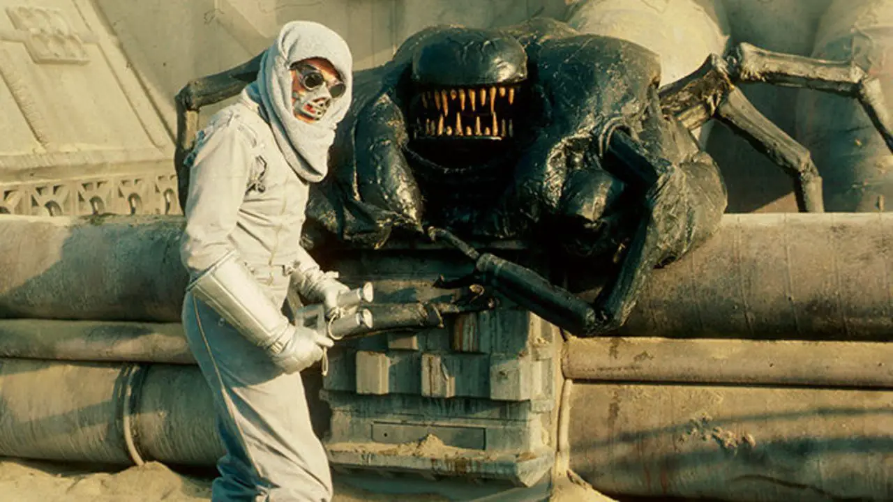 A screenshot from Forbidden World shows a man in a white desert costume complete with eye goggles, facemask and ray gun standing before a gigantic, many limbed and toothy black monster
