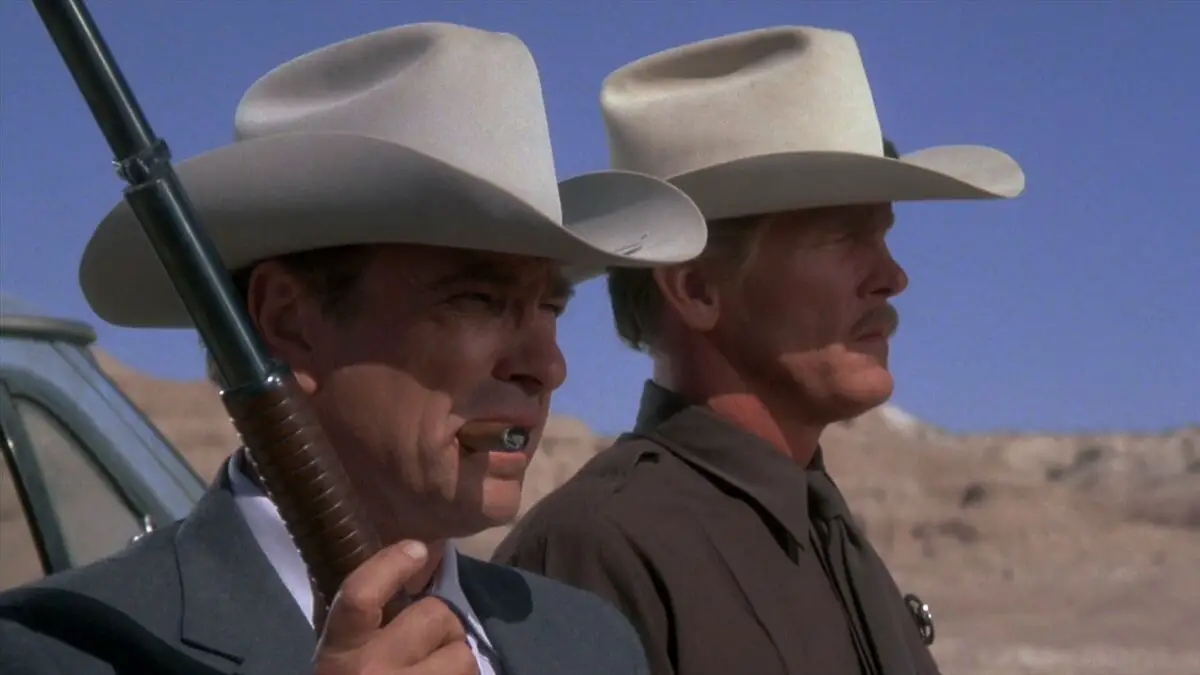 Jack and Hank stand side-by-side, Hank holding a shotgun with a cigar in his mouth.