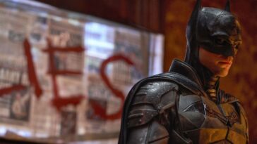 Batman stands in a room with the word "lies" drawn on the wall behind him.