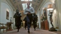 A man with a machine gun leads armed soldiers running down a grand hallway.
