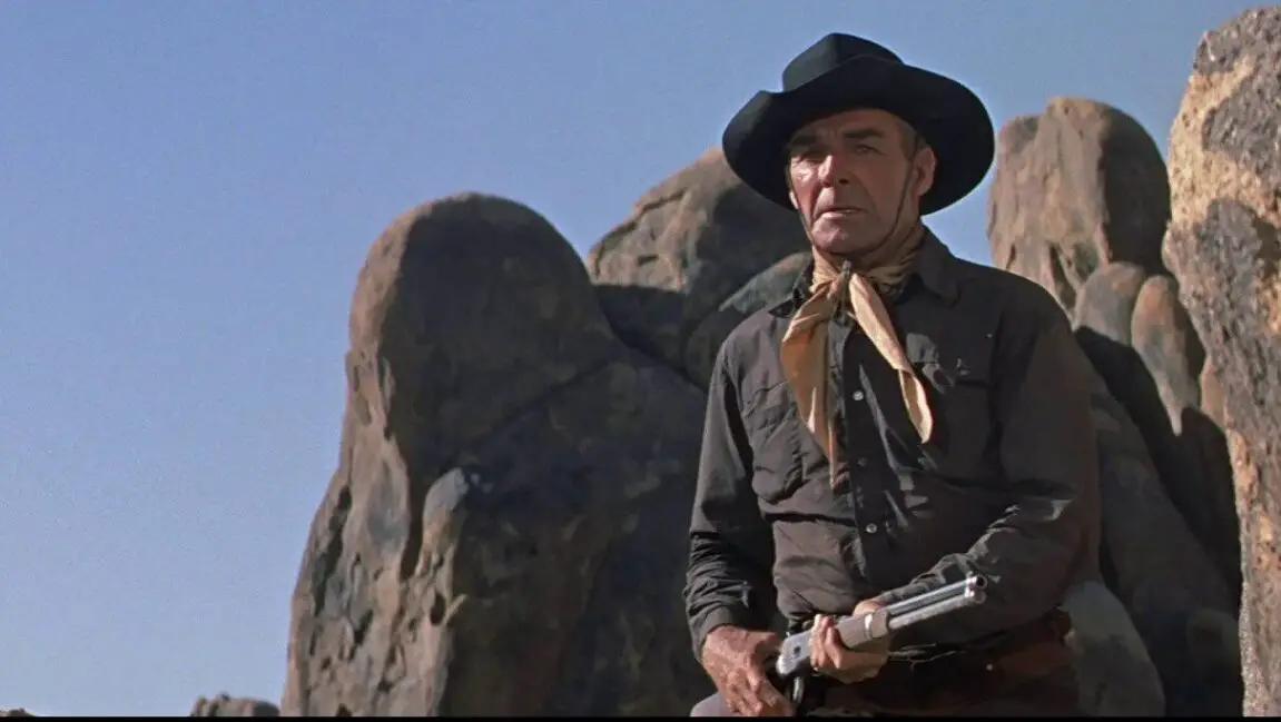 An aging cowboy stands on a rock with a shotgun in hand