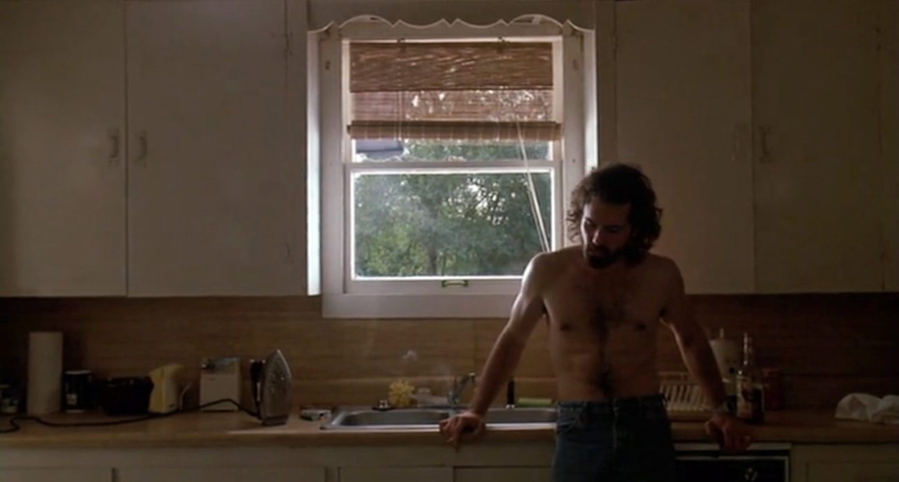A shirtless Raynor leans against a kitchen counter.