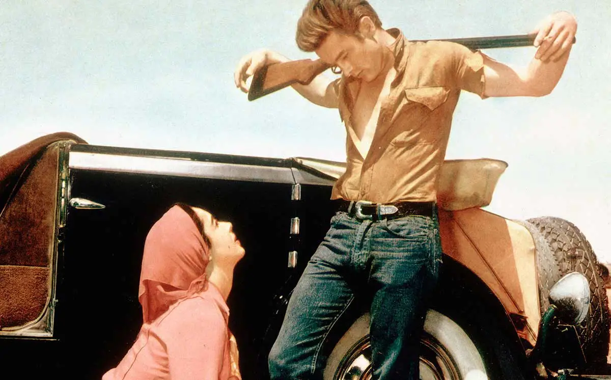 A man with a rifle over his shoulders looks down to a woman from a car fender.