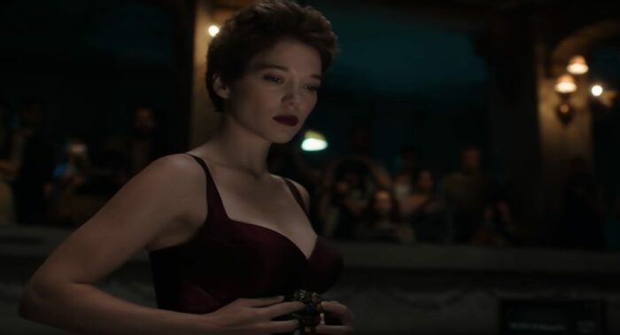Caprice (Léa Seydoux), a white woman with short, up-done brown hair, dark red lipstick, and a deep red satin bra in a dimly lit underground building. A couple of lamps on concrete columns are visible behind her. Also behind her is a crowd of people watching her perform. She is holding the remote to the surgery machine, a dark, metallic looking device against her abdomen.
