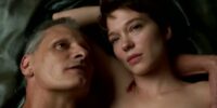 Saul (Viggo Mortensen), an older white man with short gray hair, laying with Caprice (Léa Seydoux), a white woman with short dark brown hair. They are both topless, resting atop their surgery machine while it cuts into their flesh at random. They look serene and comfortable, Saul looking off to the right, and Caprice gazing off-center. There is a fresh cut visible between her neck and shoulder as she rests her head on her forearm.