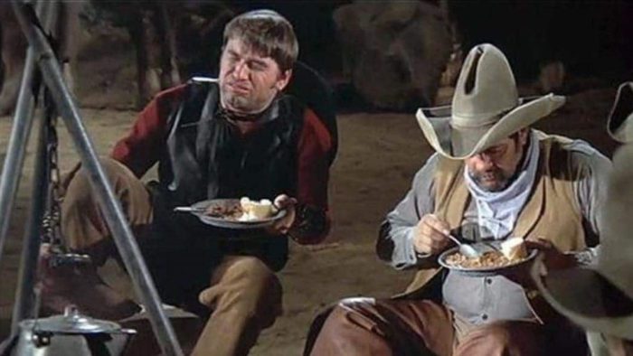 Two cowboys sitting around a campfire, enjoying a healthy diet of beans