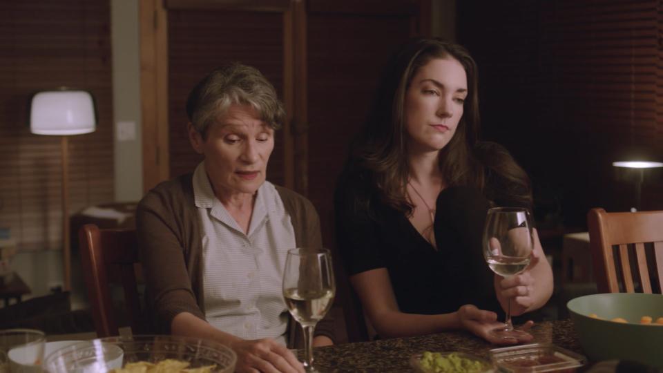 A mother and daughter talk in a kitchen over glasses of white wine.