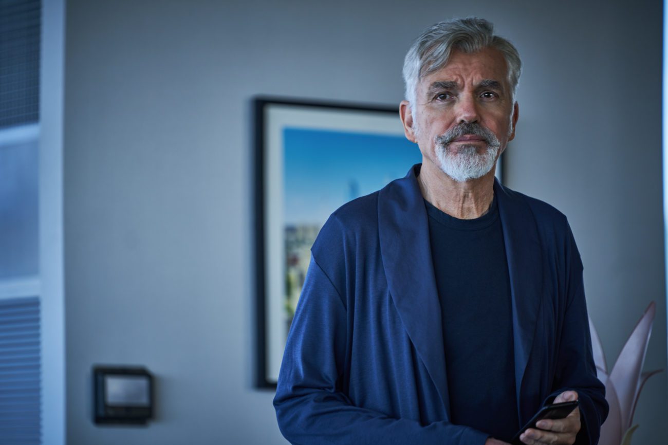 A gray-haired man stands in loungewear in his home.