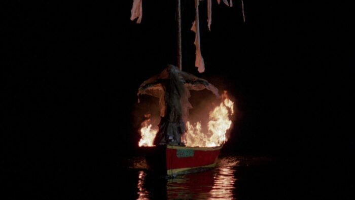 A boat on water with fire and a body on the bow.