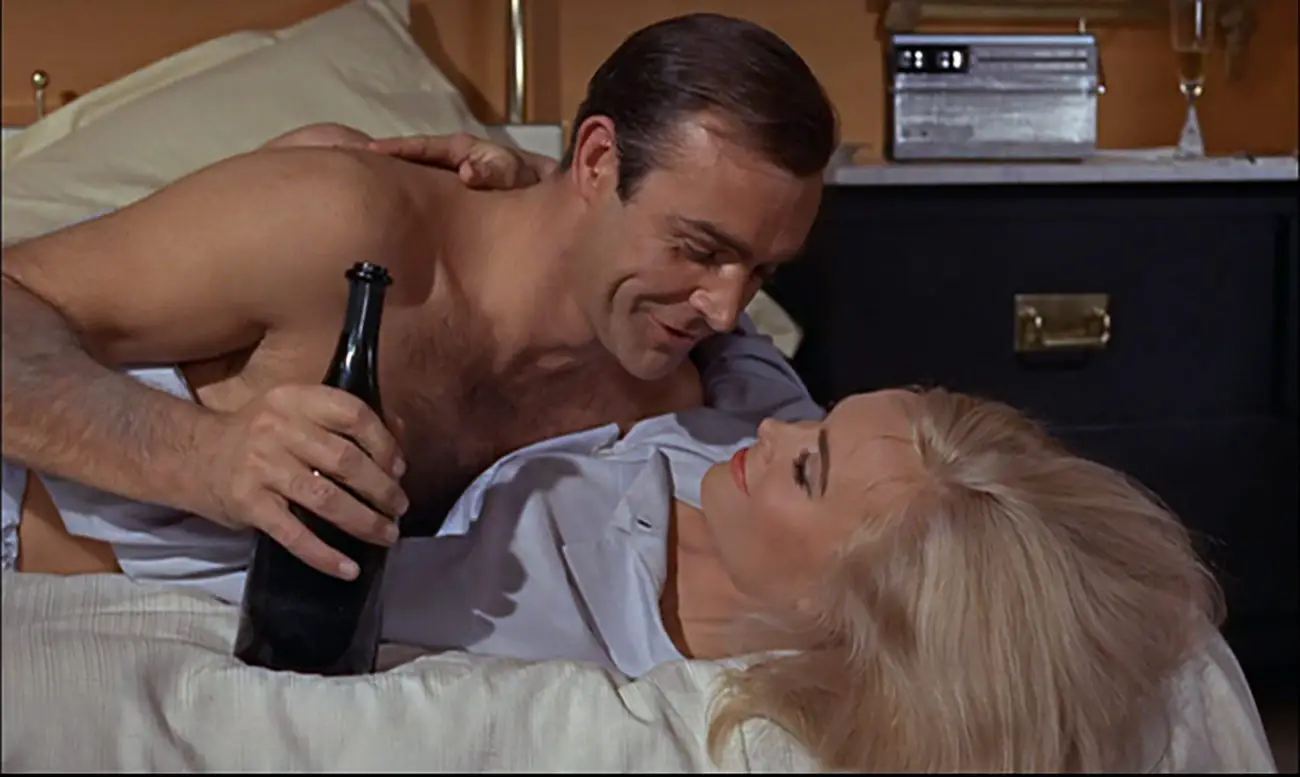 Sean Connery, holding a champagne bottle, in bed with Shirley Eaton in Goldfinger (1964)