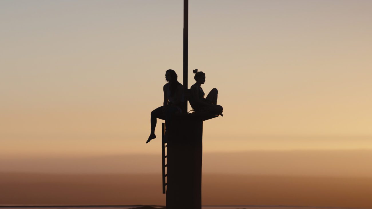 Two women sit on top of an antenna observing a sunset sky.