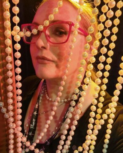 Image of Cati Glidewell, wearing red glasses and posing behind a bead curtain.