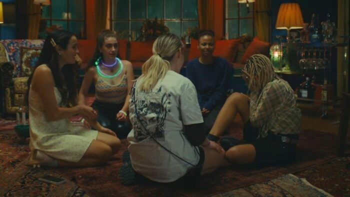 Emma, Alice (Rachel Sennott, a white girl with kinky dark brown hair wearing glowstick necklaces, a knit tank top and black leather jeans), Jordan (Myha'la Herrold, a black girl with updone brown hair wearing a dark blue sweater), Sophie, and Bee sitting in a circle inside the mansion. Rain pours outside, and the room is illuminated by various lamps and an assumed overhead light. The floor is covered in various noisily-designed rugs and the interior generally appears old-fashioned. Sophie has her face in her hands, though the energy of the picture is playful.
