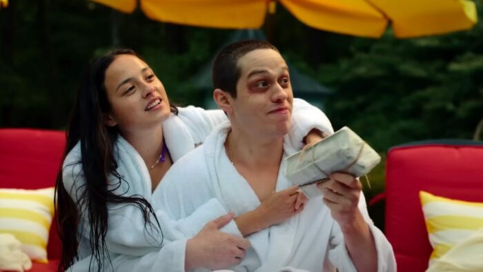 Emma (Chase Sui Wonders), a pale girl with long, straight dark hair, in a bathrobe with her hands around her boyfriend, David (Pete Davidson), a pale boy with buzzed dark brown hair and dark eyes, one of which appears to be bruised. He is also wearing a white bathrobe. He is holding a wrapped pan of zucchini bread. They are outside in overcast light, sitting on red cushioned pool chairs with yellow and white striped pillows. Forest greenery can be seen in the background along with a yellow poolside umbrella.