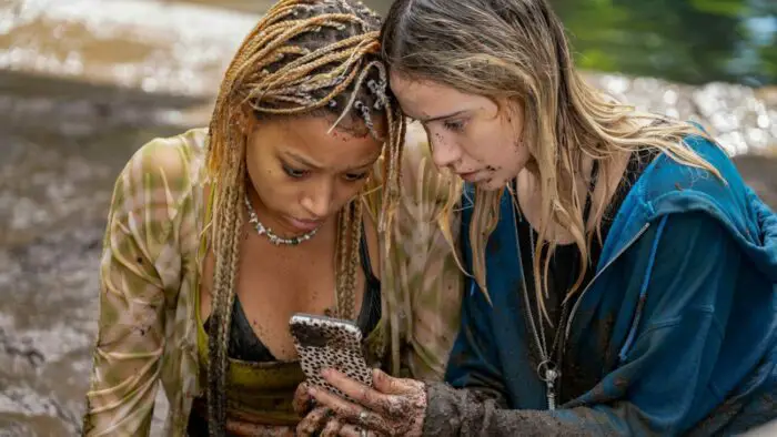 Sophie, a black girl with pale-brown skin and blonde braids, wearing a sheer green, tan, and white tie-dyed shirt, a black leather bra, moss green tank top, and shell necklace, splattered with mud, kneeling on the poolside after the storm next to her girlfriend, Bee. Bee (Maria Bakalova) is a white woman with dirty blonde hair wearing a dark blue hoodie, also splattered with mud. They are looking at a phone with a white and black spotted case.