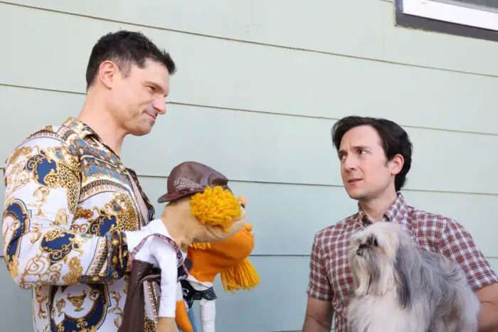 Clos (Flula Borg), a white man with very short dark brown hair wearing a fancy robe with intricate gold, white, and dark blue designs, holding two puppets on his hands talking to Sid (Josh Brener), a short white man with short dark brown hair wearing a short-sleeved gingham button-up. He is holding his pet shih-tzu in his left hand.