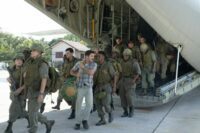 A plain clothes man with a duffel bag walks with a platoon of soldiers off a plane.