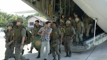 A plain clothes man with a duffel bag walks with a platoon of soldiers off a plane.