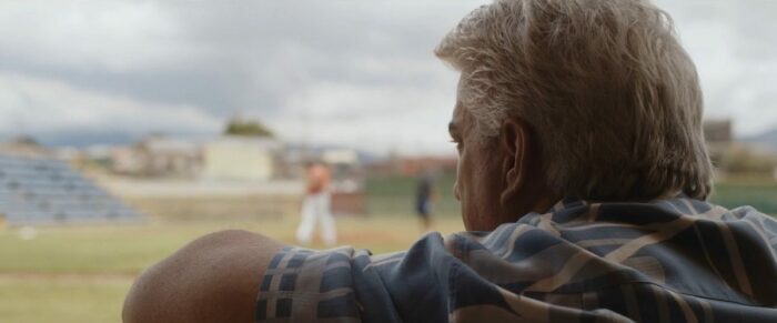 Image from The Last Out: A sliver-haired man watches a baseball pitcher in the distance. 