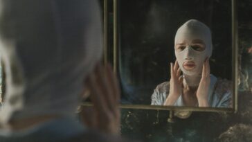 A woman in a white post-surgery mask looks at herself in a mirror